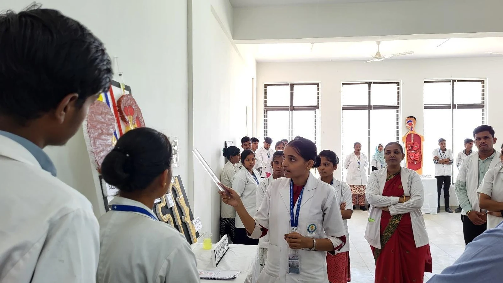BSc Nursing student from BLDEAS College of Nursing Jamkhandi presenting Group 2 Renal System anatomy model with kidney cross-section chart to judges and guests at Anatomy and Physiology Model Exhibition March 21 2025 with RENAL SYSTEM label visible on display table