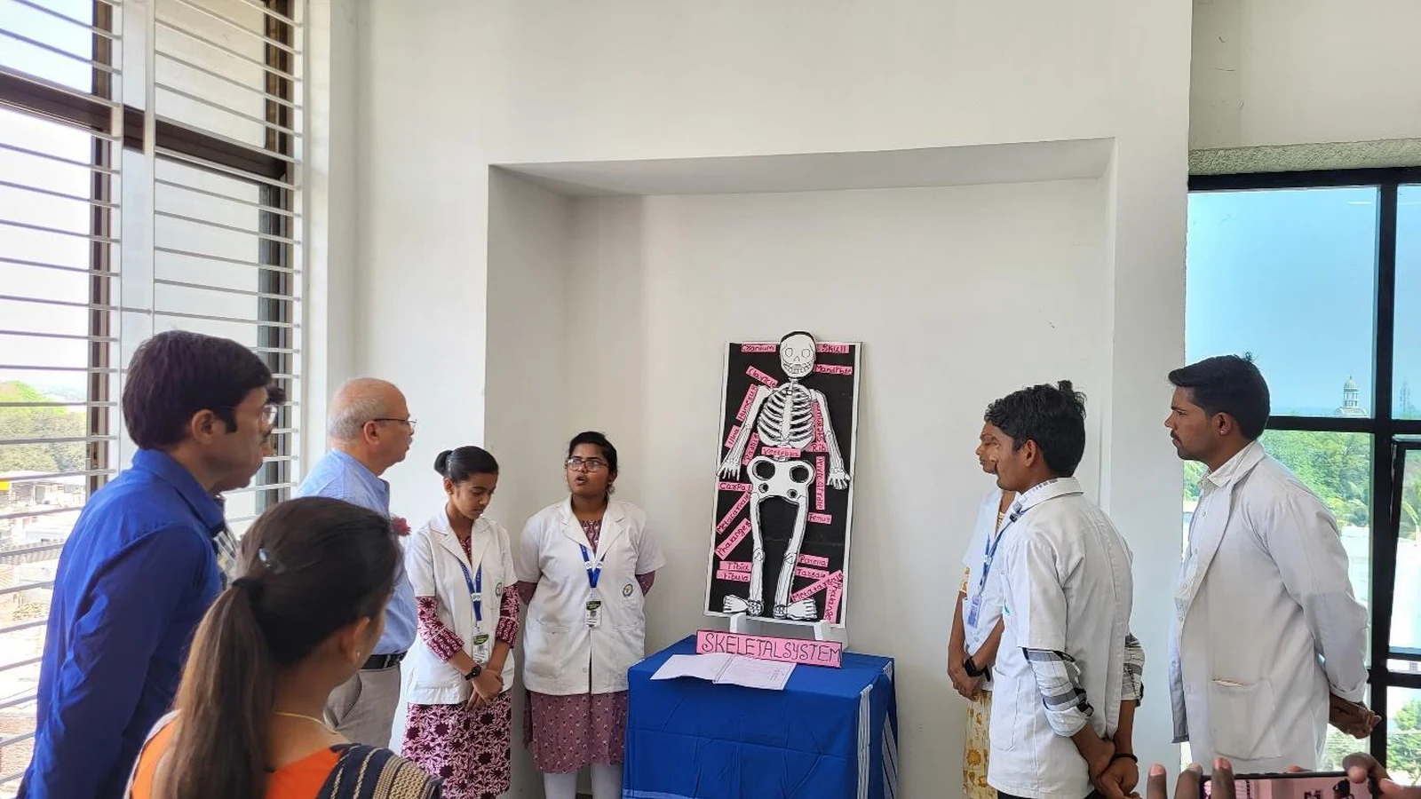 Dr Veeranna S Jatti and Dr Shankar Teli examining Group 6 Musculoskeletal Skeletal System anatomy model featuring black poster with detailed pink and white skeleton illustration at Anatomy and Physiology Model Exhibition BLDEAS College of Nursing Jamkhandi March 21 2025 with SKELETAL SYSTEM label