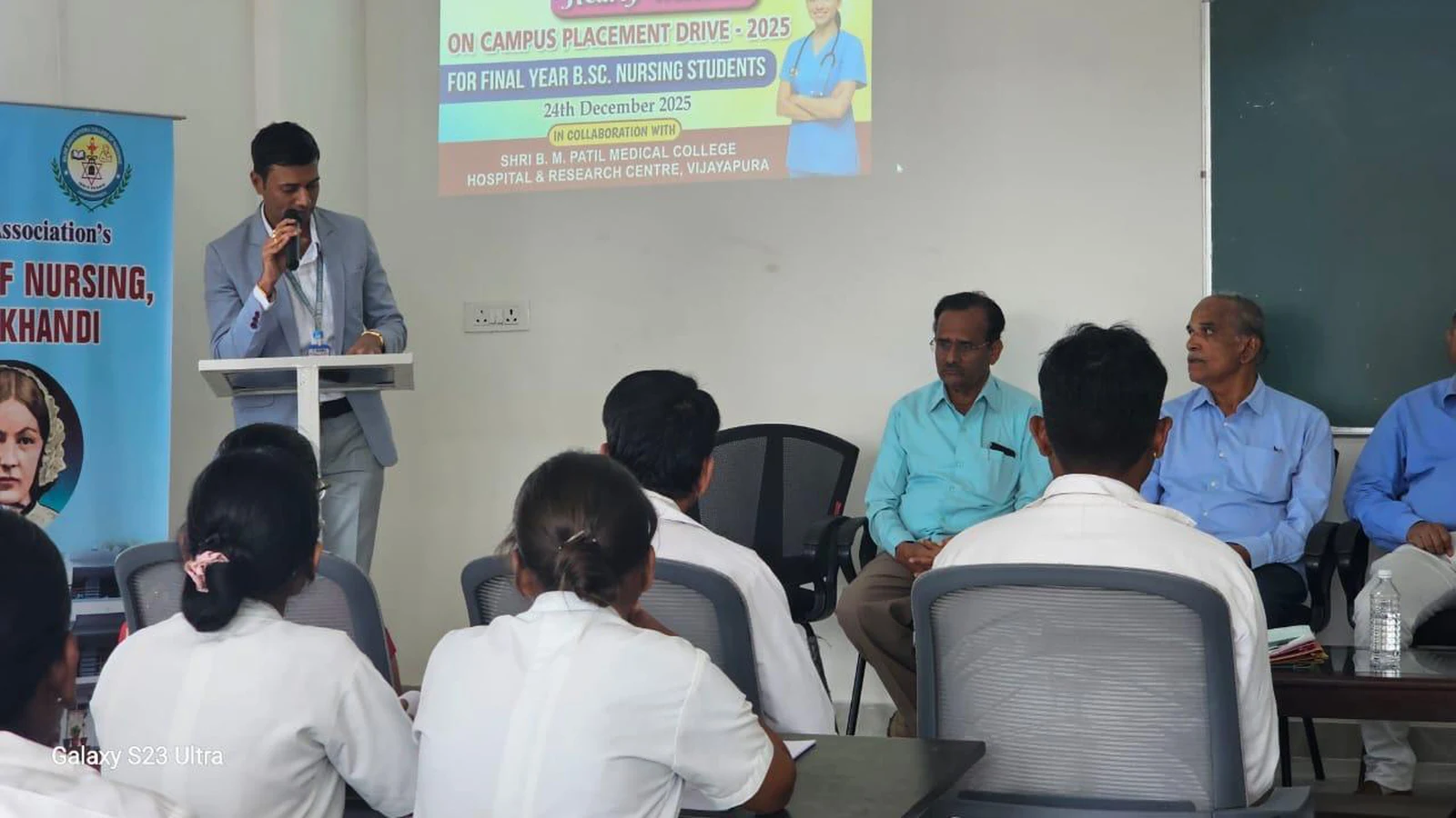 Faculty coordinator addressing final year BSc Nursing students at podium during Campus Placement Drive 2025 at BLDEA College of Nursing Jamkhandi with dignitaries and interview panel seated behind and placement drive projection visible December 2025