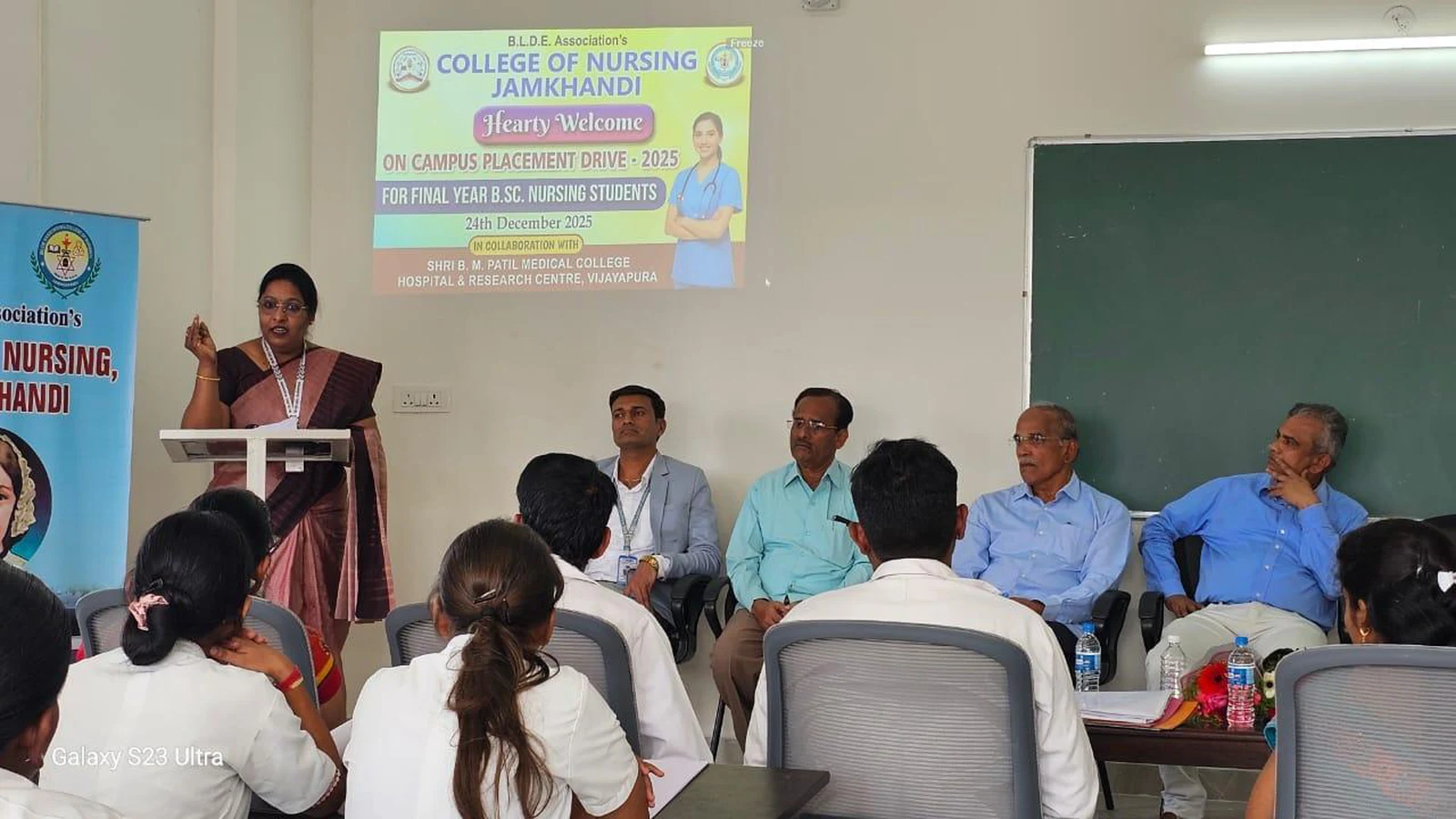 Faculty member in saree addressing final year BSc Nursing students and interview panel at podium during Campus Placement Drive 2025 at BLDEA College of Nursing Jamkhandi with placement drive banner on projection screen December 24 2025