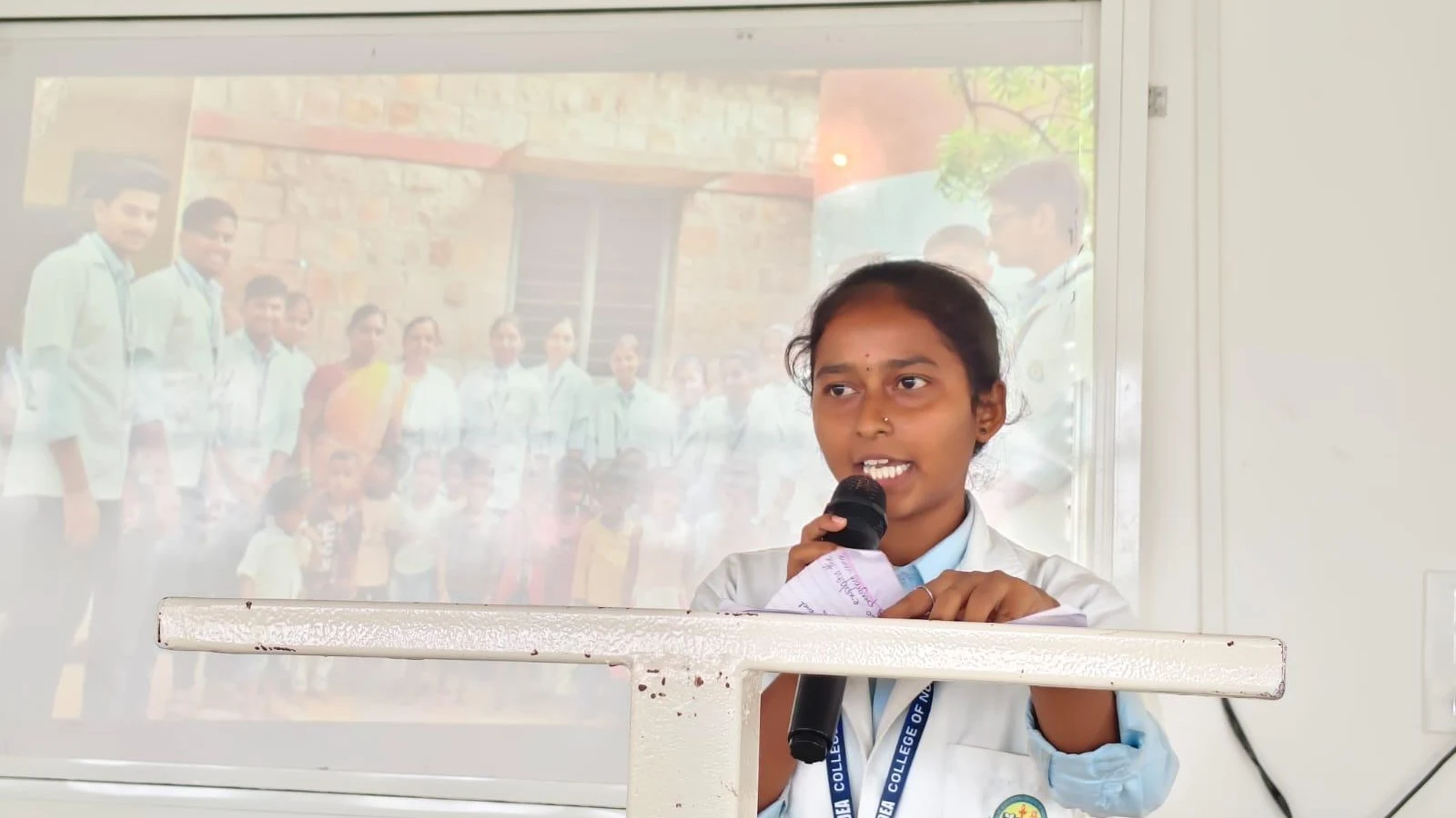 BSc Nursing student Ms Chaitra Bangalore or Ms Ayisha Hunshayal from BLDEAS College of Nursing Jamkhandi presenting welcome address at Community Health Nursing Report Presentation on 15-day Mareguddi village field posting July 19 2025 with Report Presentation projector screen visible behind podium