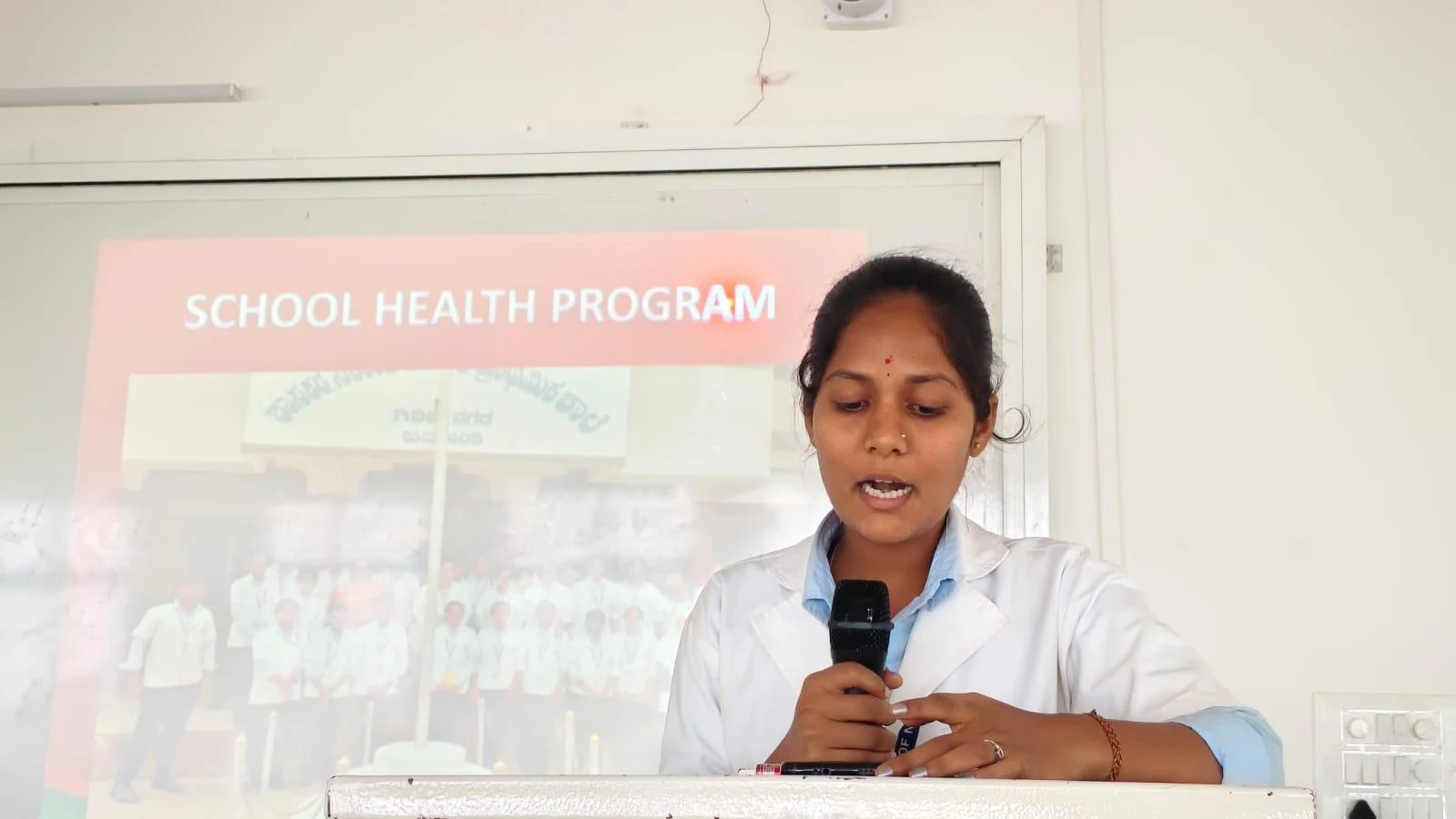 BSc Nursing student from BLDEAS College of Nursing Jamkhandi presenting School Health Program activities from 15-day Community Health Nursing posting at Mareguddi village at report presentation July 19 2025 with School Health Program projector slide visible on screen behind