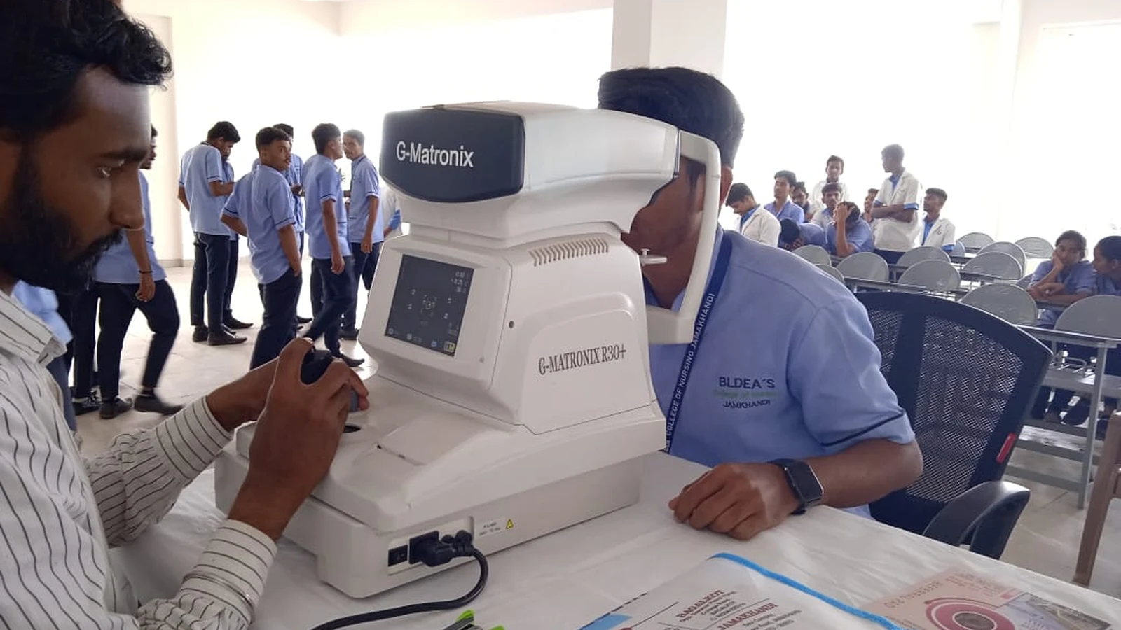 BLDEAS College of Nursing Jamkhandi student in blue nursing uniform operating G-Matronix K30 Plus auto-refractometer during Free Eye Screening Camp at Seminar Hall September 4 2024 with rows of nursing students in blue uniforms waiting for eye screening in background