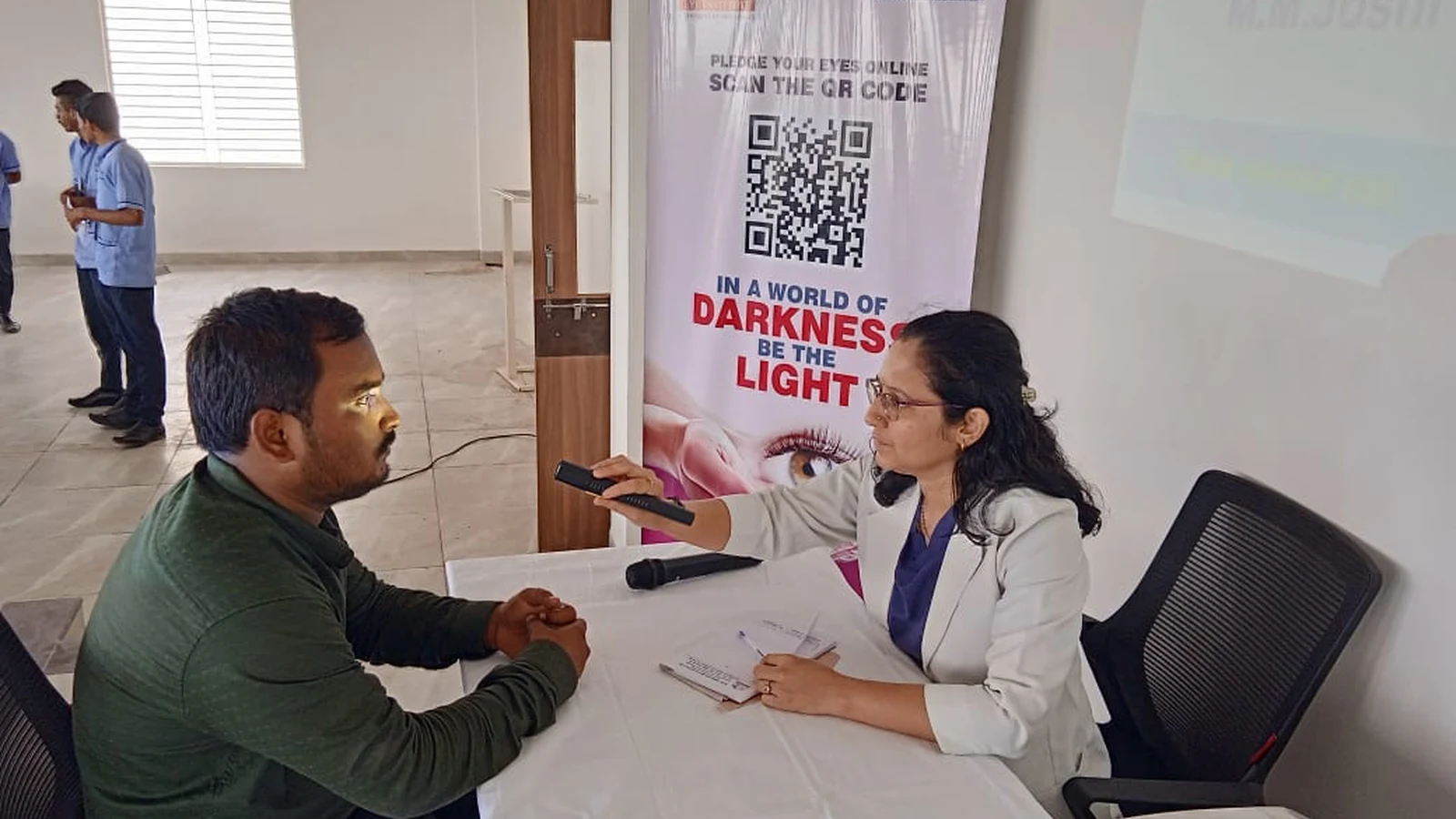 Dr Deepa Udagatti Consultant Ophthalmologist MM Joshi Eye Institute Jamkhandi using pen light torch to examine patient during Free Eye Screening Camp at Seminar Hall BLDEAS College of Nursing Jamkhandi September 4 2024 National Eye Donation Week with SGM eyebank MM Joshi Eye Institute In a World of Darkness Be the Light banner in background
