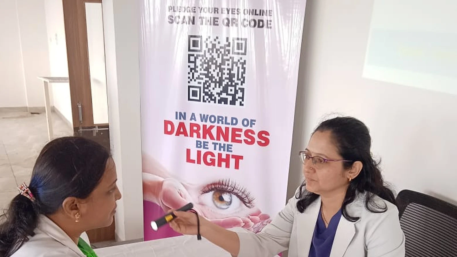 BLDEAS College of Nursing Jamkhandi faculty member in white coat receiving eye examination from Dr Deepa Udagatti Consultant Ophthalmologist MM Joshi Eye Institute Jamkhandi using pen light during Free Eye Screening Camp at Seminar Hall September 4 2024 with MM Joshi SGM eyebank Pledge Your Eyes Online In a World of Darkness Be the Light rollup banner prominently visible