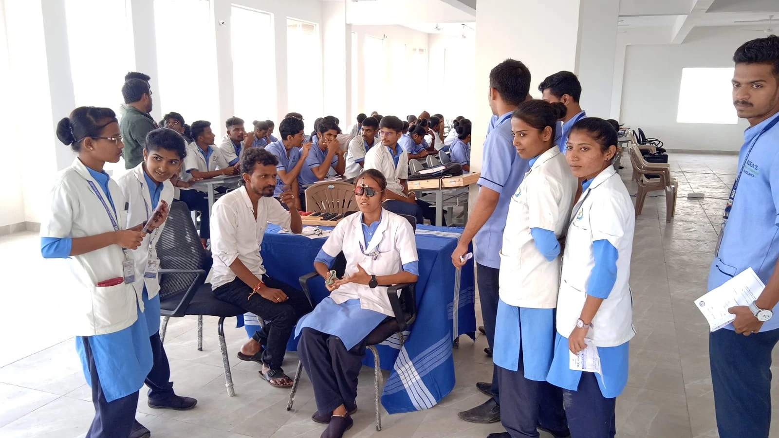 Wide view of Free Eye Screening Camp at BLDEAS College of Nursing Jamkhandi Seminar Hall September 4 2024 showing Dr Deepa Udagatti Consultant Ophthalmologist MM Joshi Eye Institute Jamkhandi seated at blue screening table surrounded by BLDEAS College of Nursing students in blue nursing uniforms attending the free eye screening camp in collaboration with MM Joshi Eye Institute Jamkhandi
