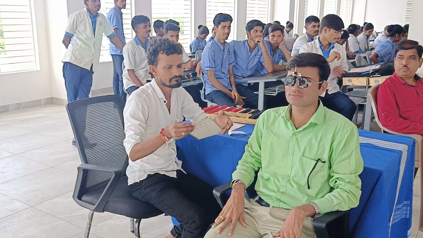 MM Joshi Eye Institute Jamkhandi optometry technician in green shirt conducting refraction test with trial frame glasses on male patient during Free Eye Screening Camp September 4 2024 at Seminar Hall BLDEAS College of Nursing Jamkhandi with rows of nursing students in blue uniforms waiting in background