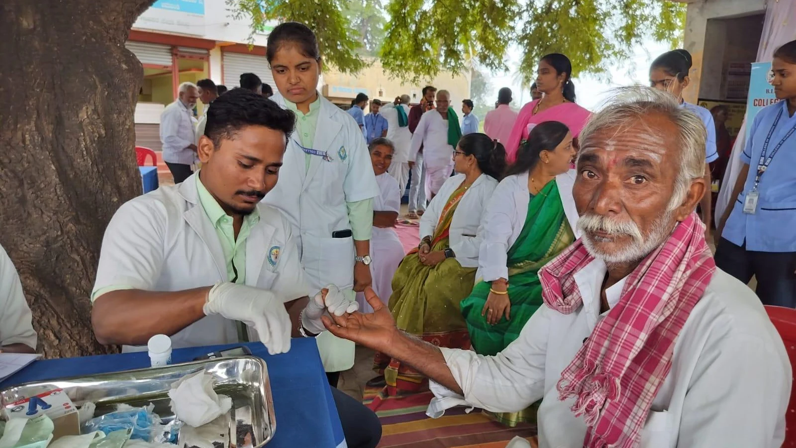 Medical student performing blood glucose finger-prick test on elderly villager seated outdoors during free health check-up camp organized by BLDEAS College of Nursing Jamkhandi at Mareguddi village Karnataka August 1 2025