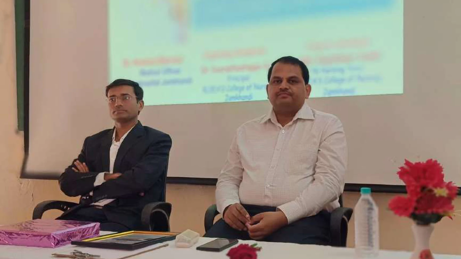 Dr Krishna Bannad Medical Officer Taluka Hospital Jamkhandi and faculty member of BLDEAS College of Nursing Jamkhandi seated at dais table during guest lecture on Bridging The Gap Cancer Awareness Early Detection and Patient Support at Lecture Hall No 1 BLDEAS College of Nursing Jamkhandi November 9 2024 with event program projected on screen in background