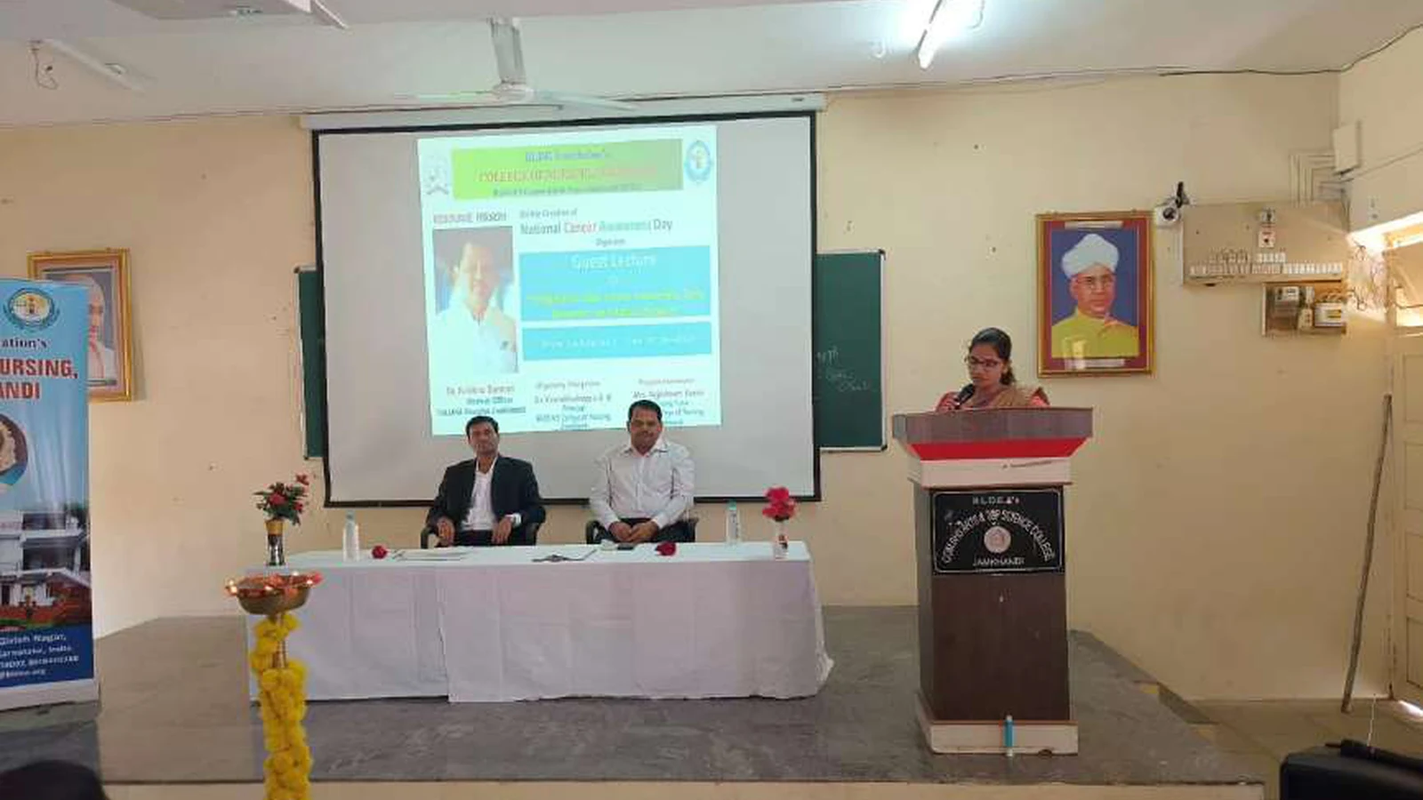 Mrs Rajeshwari Kambi PG Nursing Faculty BLDEAS College of Nursing Jamkhandi delivering welcome address at podium during guest lecture on Bridging The Gap Cancer Awareness Early Detection and Patient Support at Lecture Hall No 1 BLDEAS College of Nursing Jamkhandi November 9 2024 with dignitaries seated at dais table and event screen visible in background