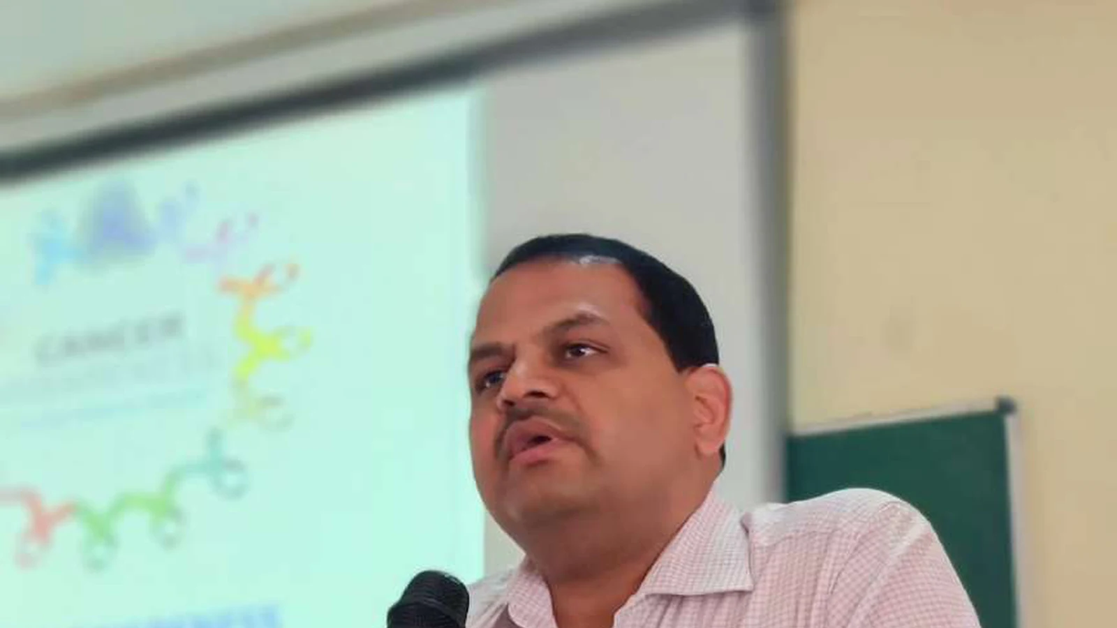 Dr Krishna Bannad Medical Officer Taluka Hospital Jamkhandi delivering guest lecture on cancer awareness at podium with microphone at Lecture Hall No 1 BLDEAS College of Nursing Jamkhandi November 9 2024 with cancer awareness educational slide projected on screen behind him