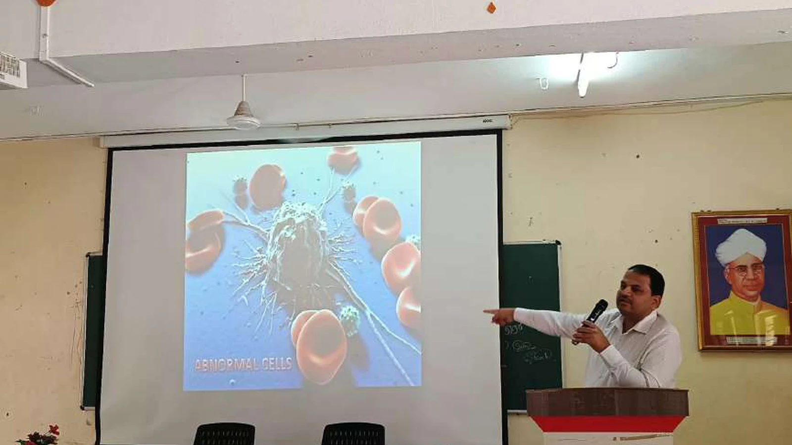 Dr Krishna Bannad Medical Officer Taluka Hospital Jamkhandi pointing to projected slide showing abnormal cancer cells during guest lecture on Bridging The Gap Cancer Awareness Early Detection and Patient Support at Lecture Hall No 1 BLDEAS College of Nursing Jamkhandi November 9 2024 with BLDEAS College of Nursing podium visible