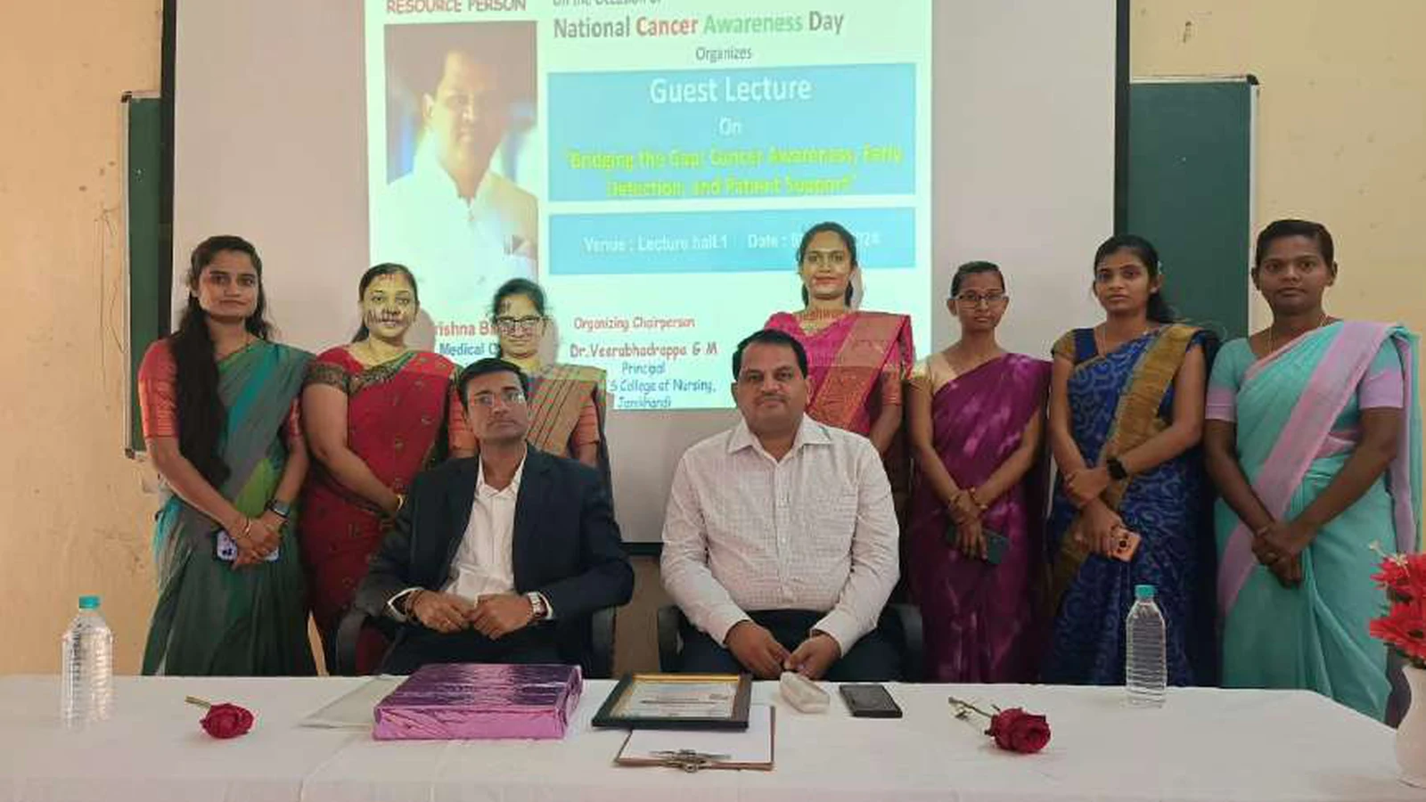 Dr Krishna Bannad Medical Officer Taluka Hospital Jamkhandi and Dr Veerabhadrappa G Mendagudli Principal BLDEAS College of Nursing Jamkhandi seated at dais table with faculty members of BLDEAS College of Nursing Jamkhandi standing for group photograph at conclusion of guest lecture on Bridging The Gap Cancer Awareness Early Detection and Patient Support at BLDEAS College of Nursing Jamkhandi November 9 2024 with event program projected on screen in background