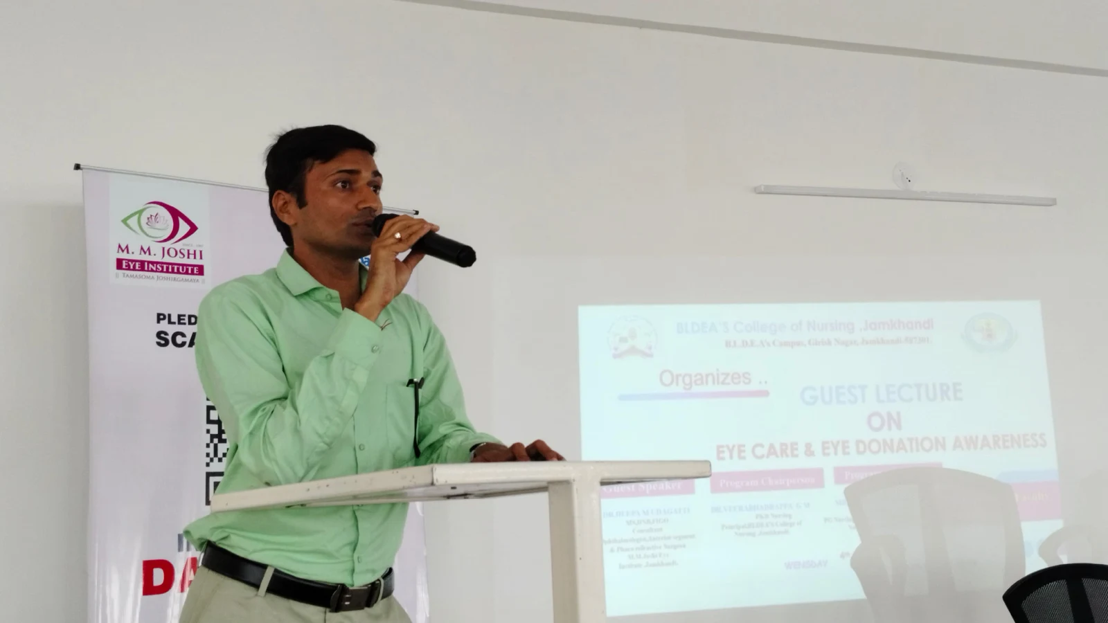 Mr Somesh Nikkam PG Nursing Tutor BLDEAS College of Nursing Jamkhandi speaking at podium during Guest Lecture on Eye Care and Eye Donation Awareness September 4 2024 with Guest Lecture on Eye Care and Eye Donation Awareness program slide projected on screen and MM Joshi Eye Institute rollup banner visible