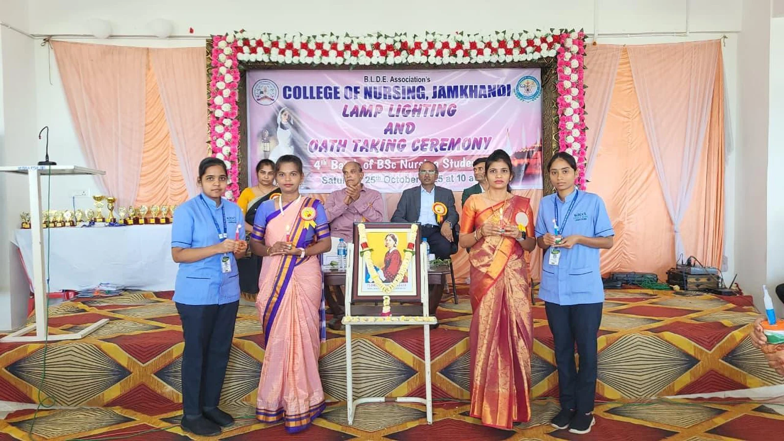 4th Batch 1st Year BSc Nursing students in blue uniform receiving light from nursing faculty leaders in procession during Lamp Lighting Ceremony at BLDEA College of Nursing Jamkhandi with ceremony banner in background October 25 2025