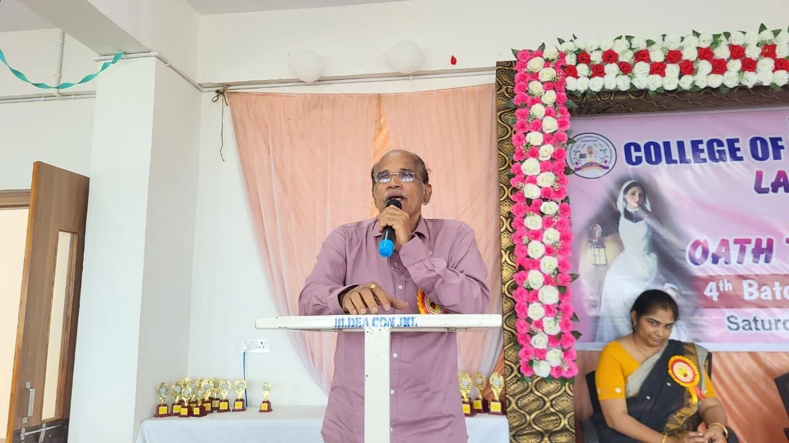 Inaugurator Dr Raveendra Madraki Chairman and Chief Nephrologist Yashoda Institute of Medical Sciences Vijayapura delivering inaugural address at podium during Lamp Lighting and Oath Taking Ceremony 2025 at BLDEA College of Nursing Jamkhandi with banner and trophies visible
