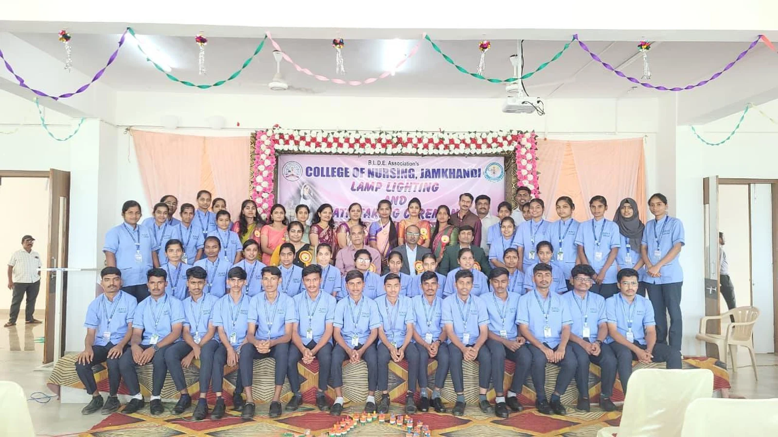 4th Batch 1st Year BSc Nursing students in blue uniform with faculty and dignitaries in group photograph at Lamp Lighting and Oath Taking Ceremony 2025 BLDEA College of Nursing Jamkhandi with ceremony banner October 25 2025
