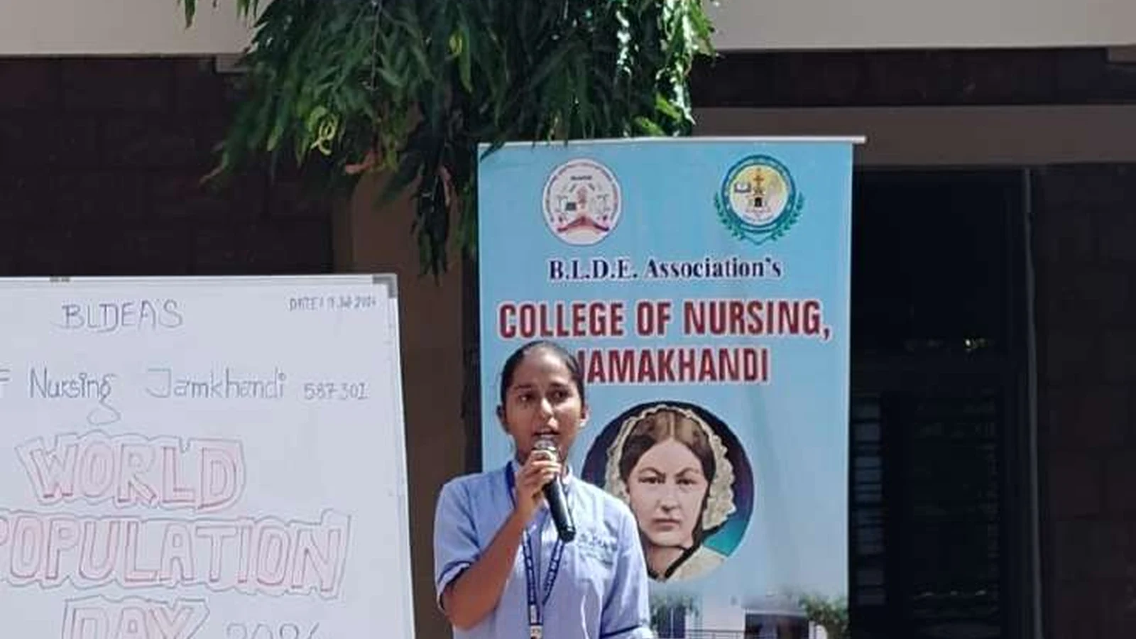 Second semester BSc Nursing student giving introduction at microphone before World Population Day 2024 mime show on college premises with World Population Day 2024 display board and BLDEAS College of Nursing Jamkhandi rollup banner in background July 11 2024