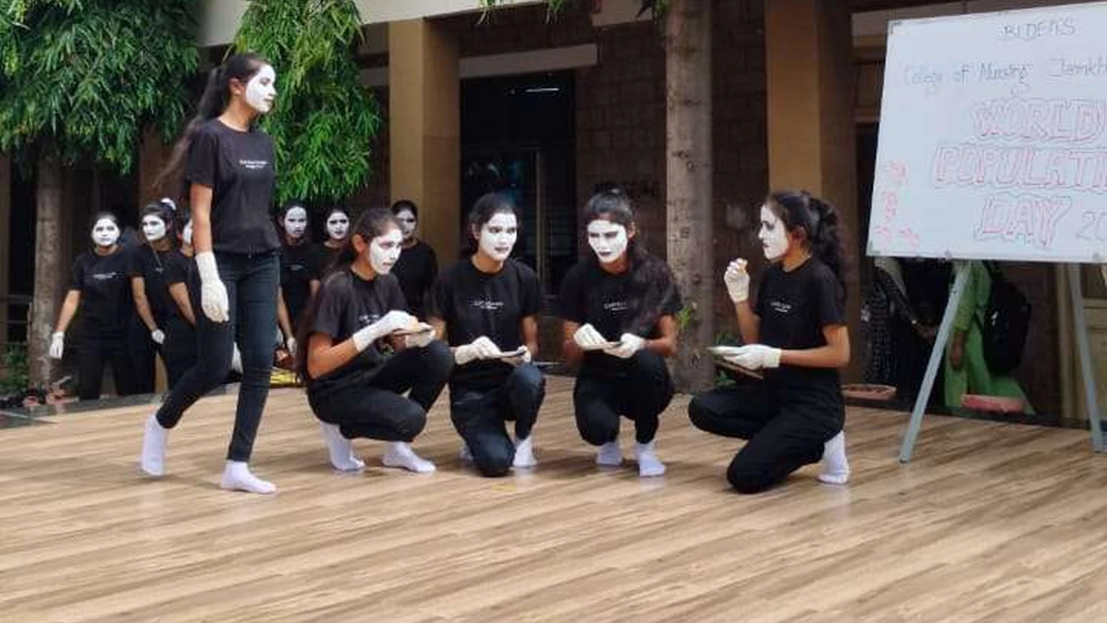 Second semester BSc Nursing students from BLDEAS College of Nursing Jamkhandi performing crouching mime scene depicting overpopulation awareness on wooden stage at college campus with Arts Block building visible in background July 11 2024