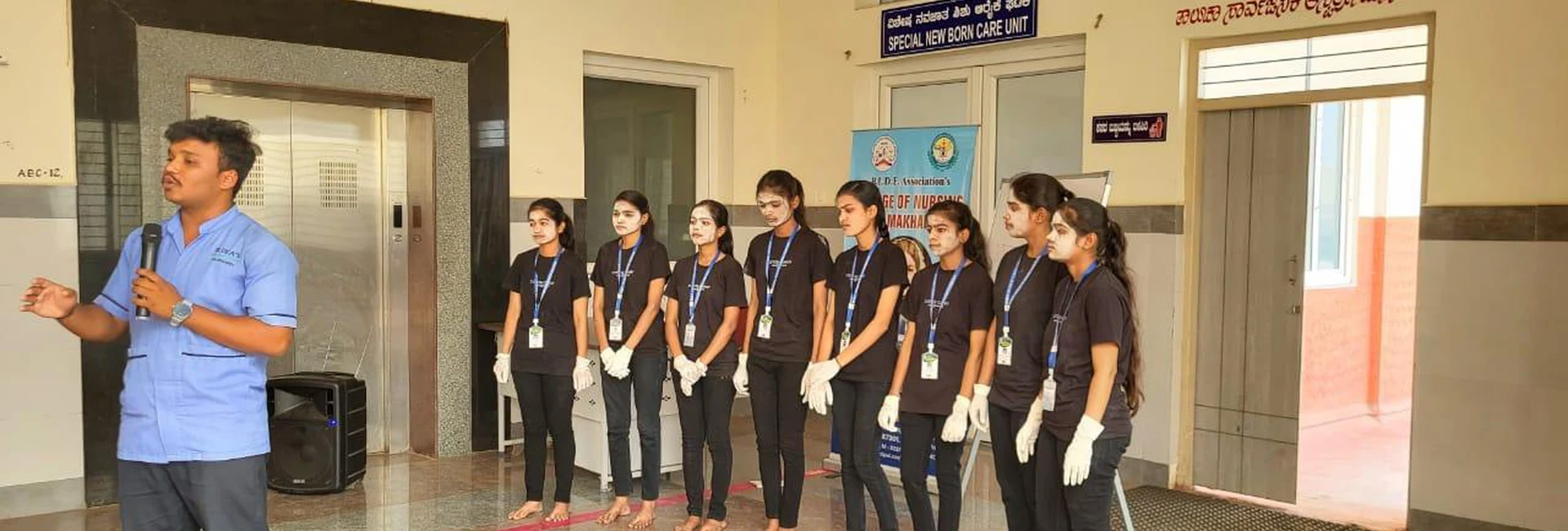 Mr Rakesh Patil third semester BSc Nursing student giving welcome speech at microphone with nursing students in black outfits lined up at OPD of Taluka Hospital Jamkhandi during World Tuberculosis Day 2025 mime show organized by BLDEAS College of Nursing Jamkhandi March 25 2025