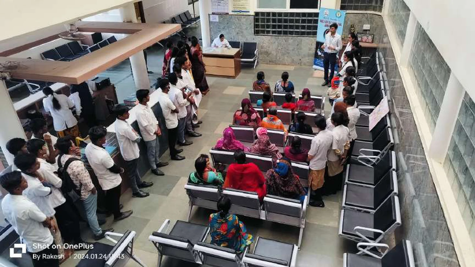 Aerial overhead view of Tungal Hospital waiting area showing first semester BSc Nursing students from BLDEAS College of Nursing Jamkhandi performing National Girl Child Day awareness skit before ANC and PNC mothers seated in chairs January 24 2024