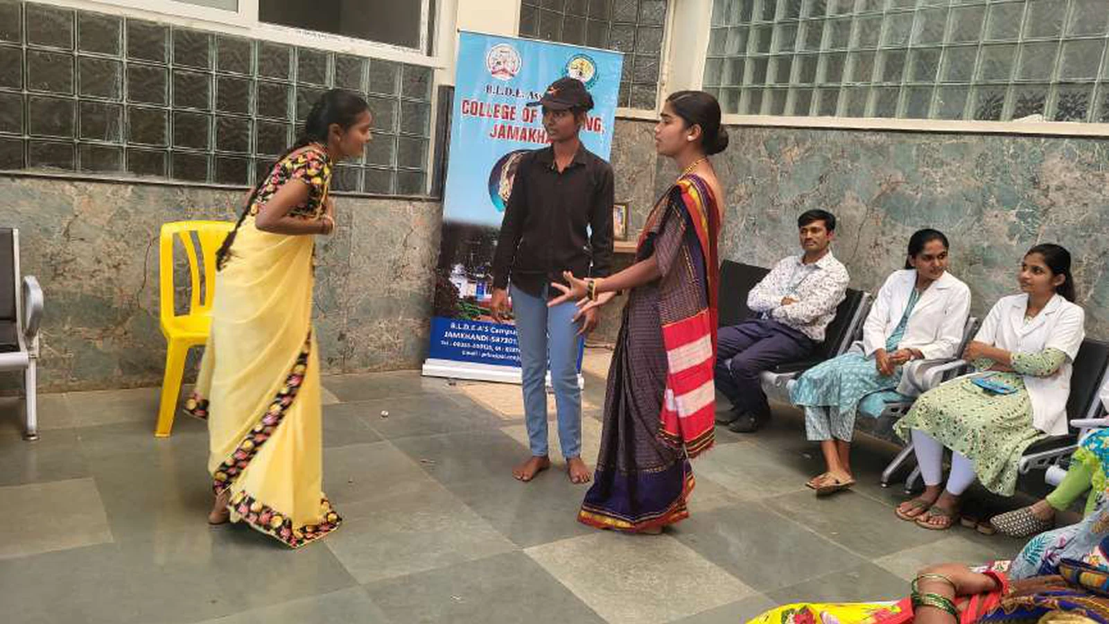 Two first semester BSc Nursing students acting in National Girl Child Day awareness skit one in yellow saree and one in striped saree at Tungal Hospital Jamkhandi with BLDEAS College of Nursing rollup banner and audience visible January 24 2024