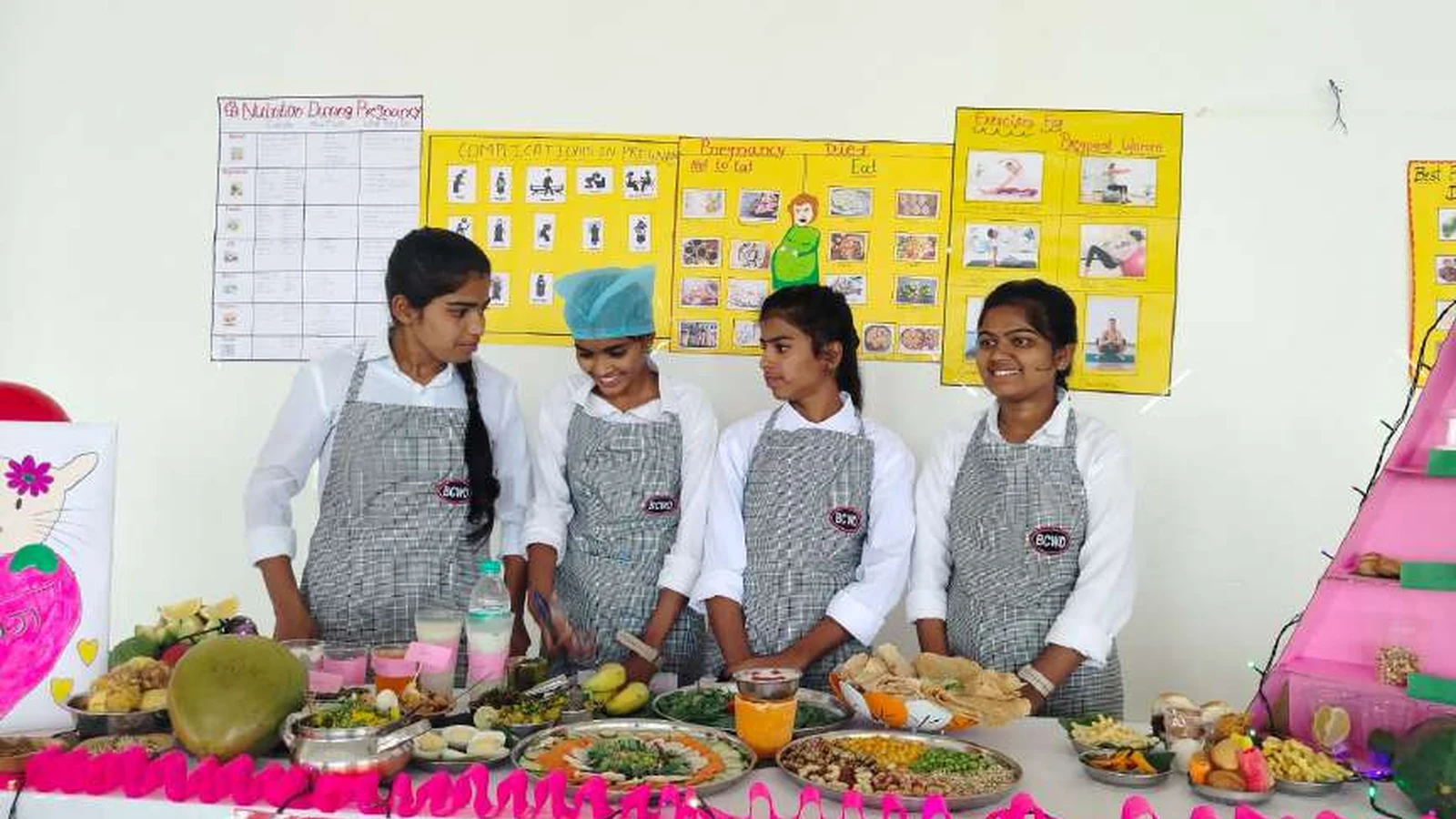 Four second semester BSc Nursing students of BLDEAs College of Nursing Jamkhandi in grey checker aprons standing at their Nutrition During Pregnancy diet exhibition stall with fruits vegetables and food items on display table and Nutrition During Pregnancy diet charts and posters on wall during Nutrition Exhibition and Demonstration Programme 2024 at Nutrition Lab BLDEAs College of Nursing Jamkhandi September 11 2024