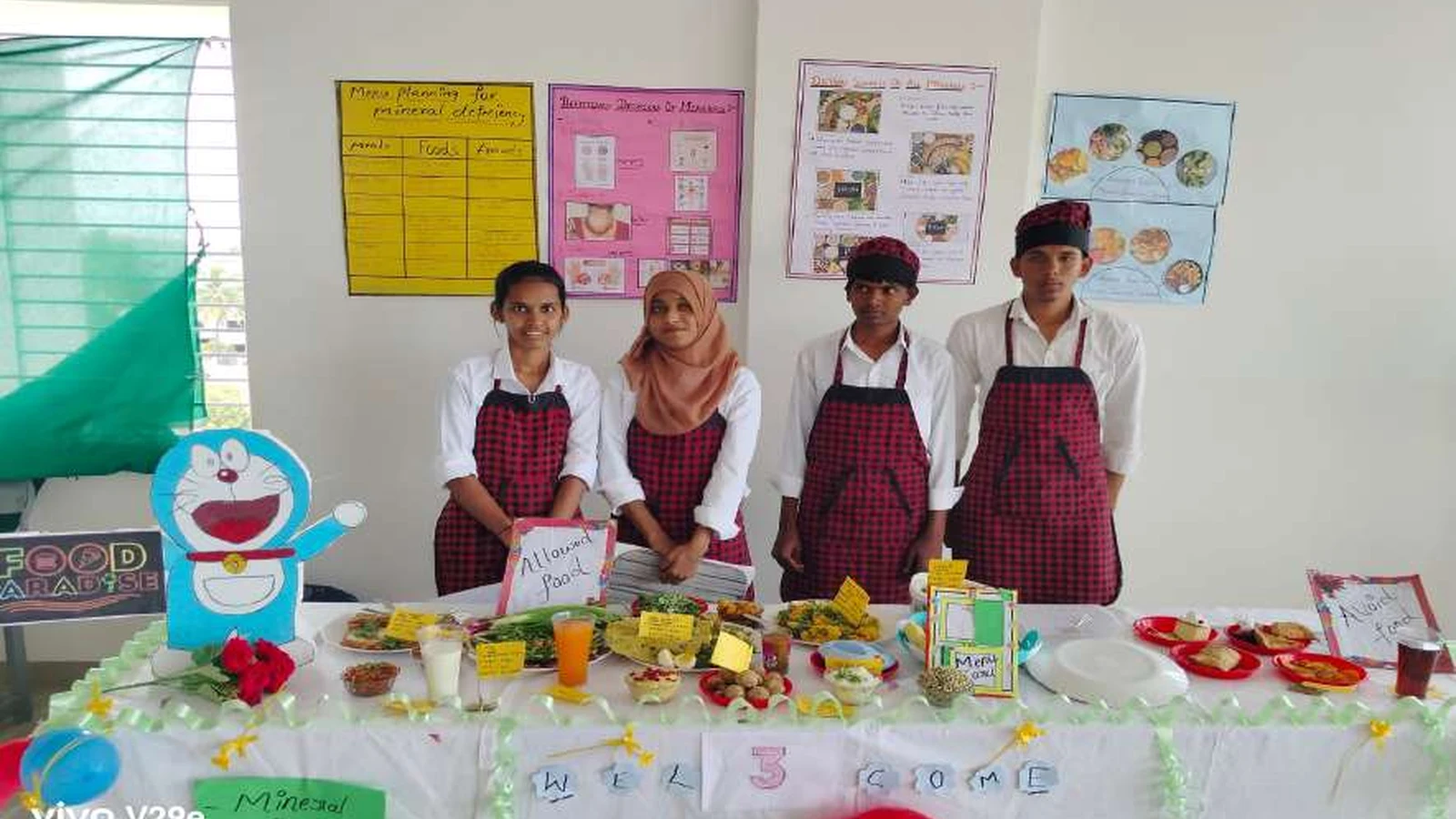 Second semester BSc Nursing students of BLDEAs College of Nursing Jamkhandi in red checked aprons at their diet for mineral deficiencies exhibition stall Food Paradise group with Doraemon mascot Welcome on tablecloth Allowed Food sign and nutrition diet charts on wall during Nutrition Exhibition and Demonstration Programme 2024 at Nutrition Lab BLDEAs College of Nursing Jamkhandi September 11 2024