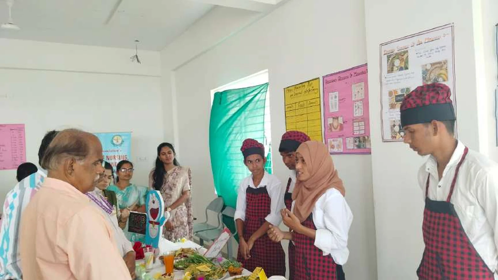 Faculty judge evaluating the diet for mineral deficiencies exhibition stall Food Paradise with second semester BSc Nursing students of BLDEAs College of Nursing Jamkhandi in red checked aprons presenting their diet plan and food items during judging session at Nutrition Exhibition and Demonstration Programme 2024 at Nutrition Lab BLDEAs College of Nursing Jamkhandi September 11 2024