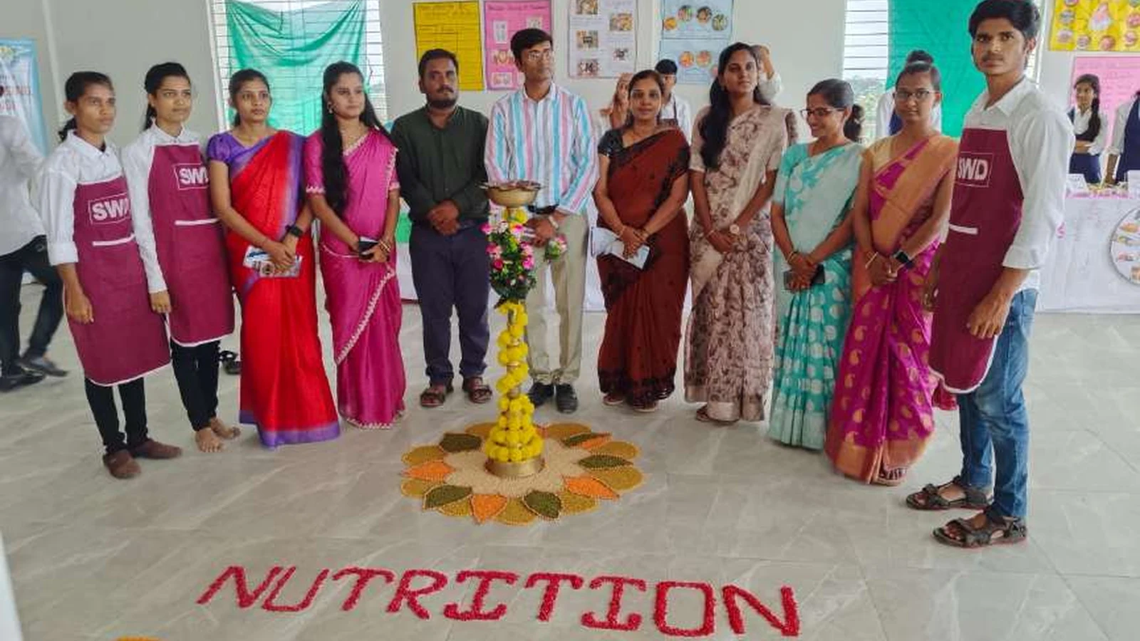 Group photograph of second semester BSc Nursing student exhibitors in SWD aprons and faculty of BLDEAs College of Nursing Jamkhandi with dignitaries at Nutrition Exhibition and Demonstration Programme 2024 at Nutrition Lab BLDEAs College of Nursing Jamkhandi September 11 2024 with NUTRITION EXHIBITION text made from seeds and spices and decorative lamp rangoli visible on floor