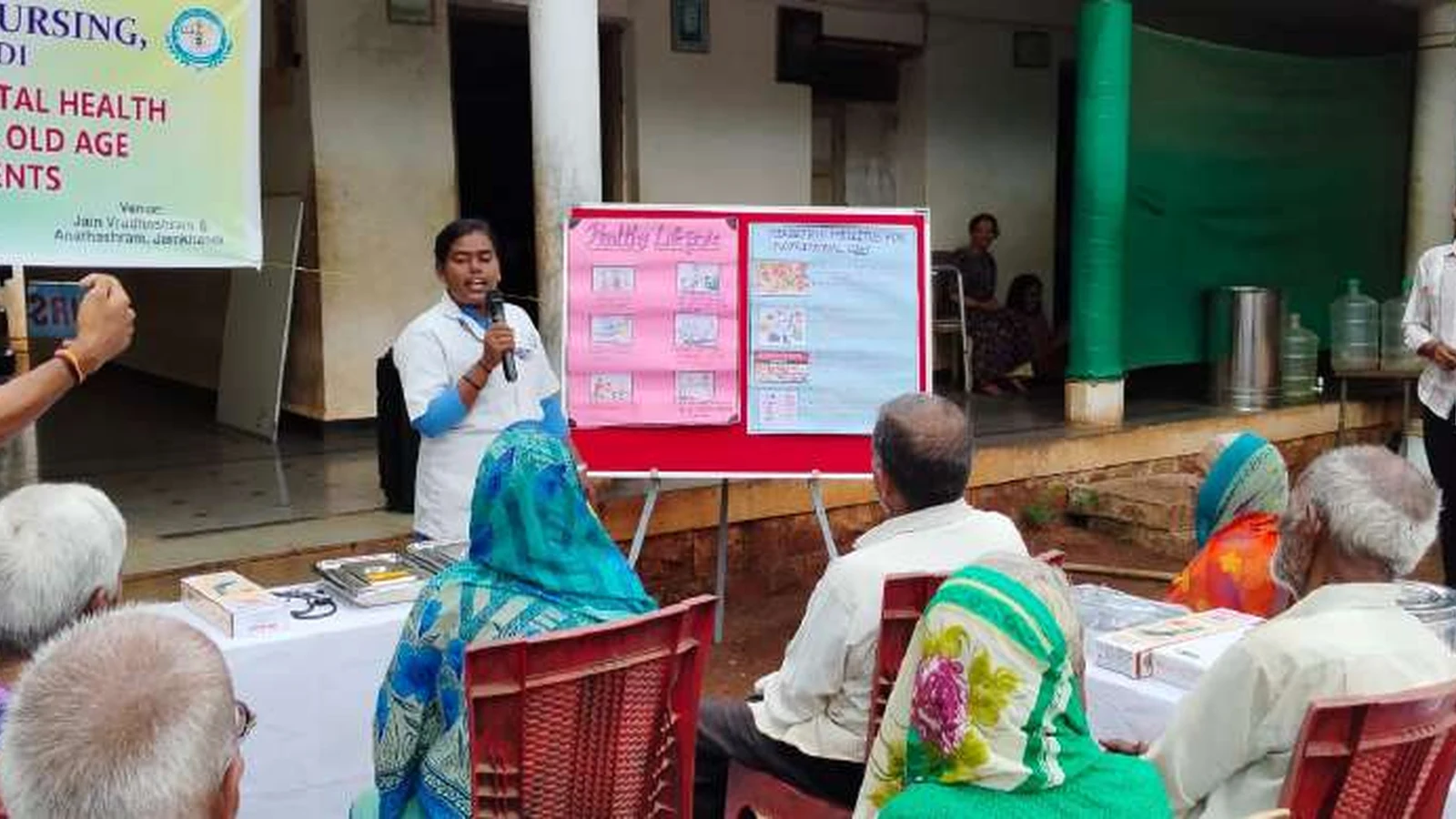 BSc Nursing student giving health education talk to elderly residents of Jain Vrudhrashram Jamkhandi on healthy lifestyle hypertension diabetes prevention and stress management with BLDEAS Geriatric and Mental Health Assessment banner and health chart visible July 18 2024