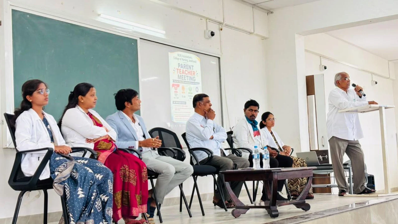 Wide view of Parent-Teacher Meeting inaugural session at BLDEA College of Nursing Jamkhandi with panel of dignitaries seated on stage and speaker addressing parents students and faculty at podium June 2026