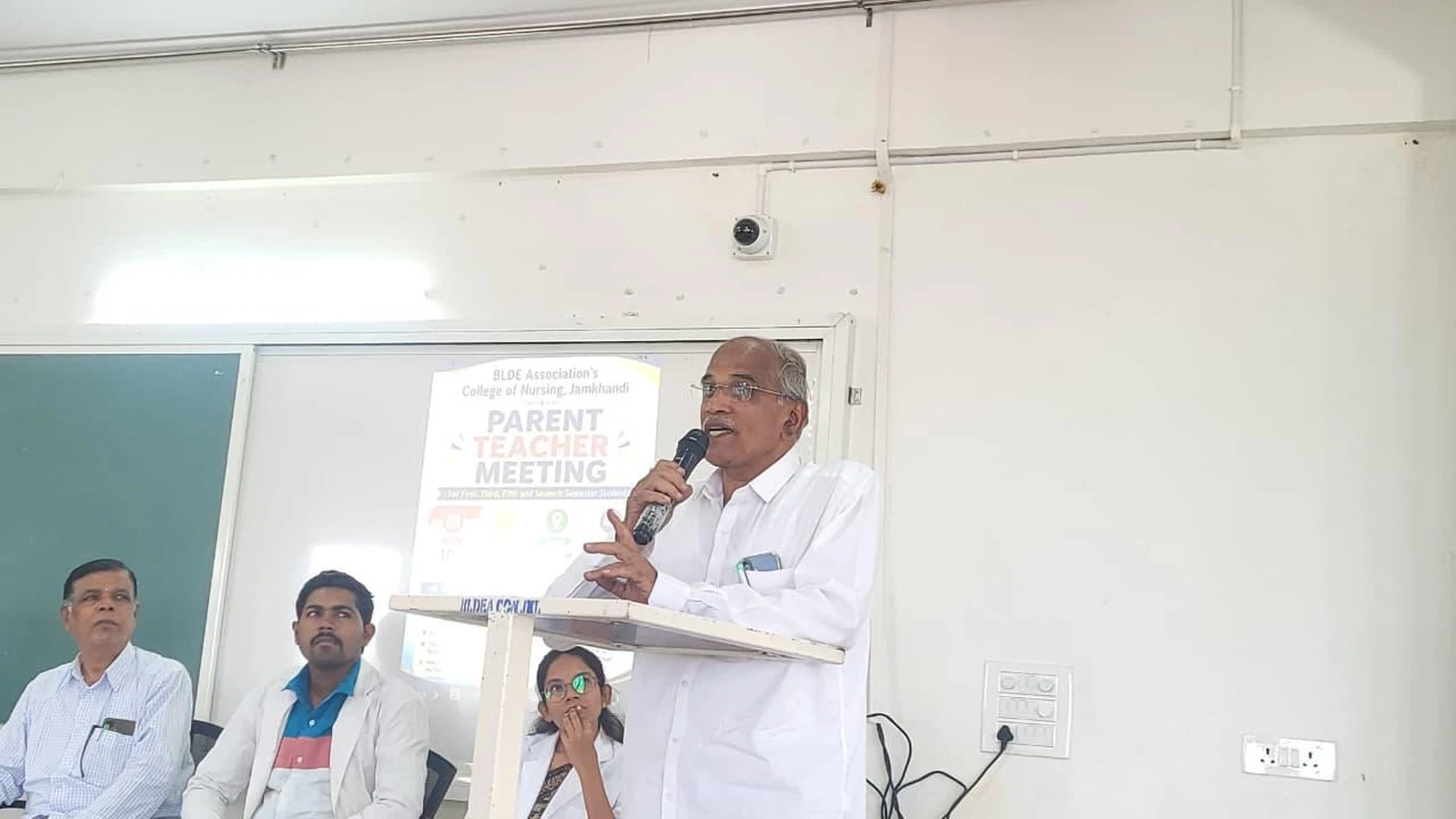 Prof S H Lagali Administrative Officer BLDE Association addressing parents and students with microphone at Parent-Teacher Meeting BLDEA College of Nursing Jamkhandi at podium with panel members seated behind June 2026