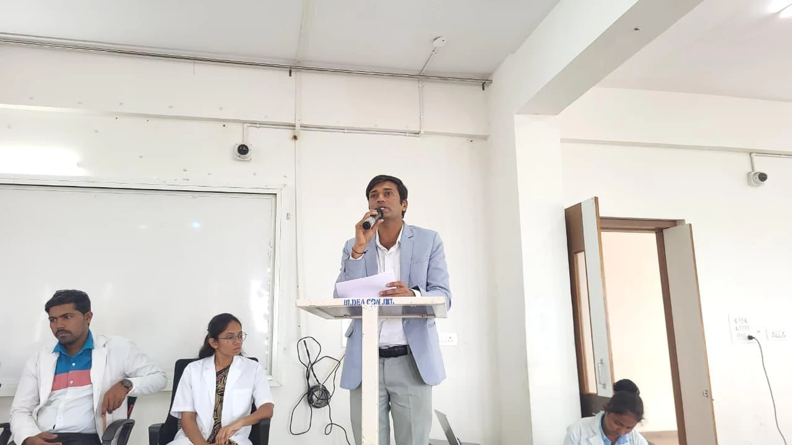 Faculty coordinator in blazer presenting to Parent-Teacher Meeting gathering at podium at BLDEA College of Nursing Jamkhandi June 2026 with colleagues seated alongside