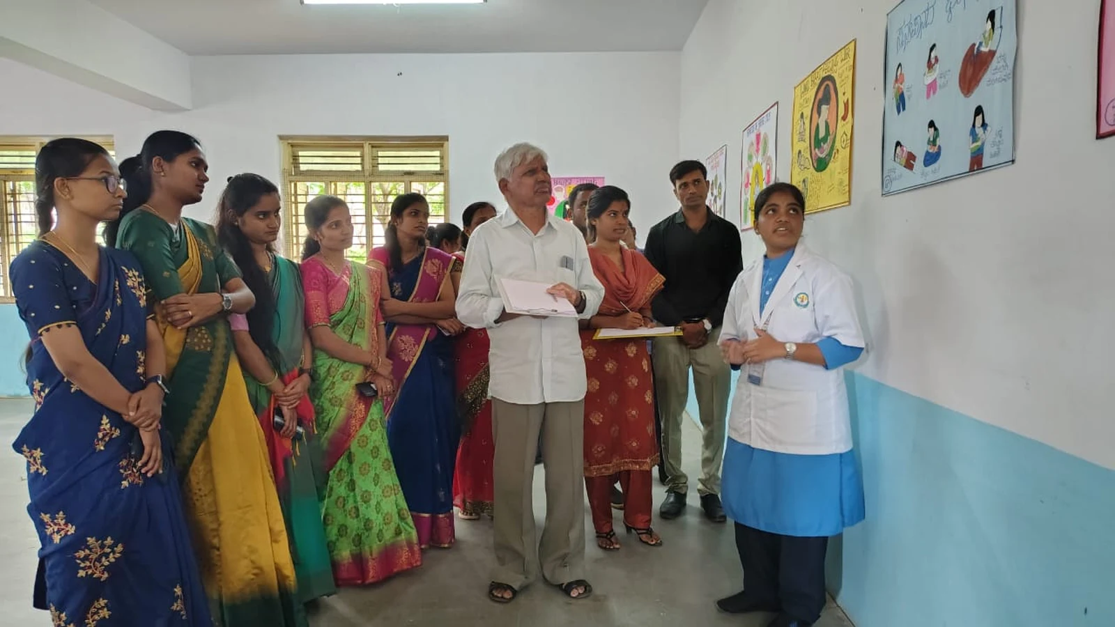 Dr H G Daddi judge with clipboard and nursing student in white coat presenting breastfeeding poster wide group of students and faculty at World Breastfeeding Week Poster Competition BLDEAS College of Nursing Jamkhandi August 7 2024