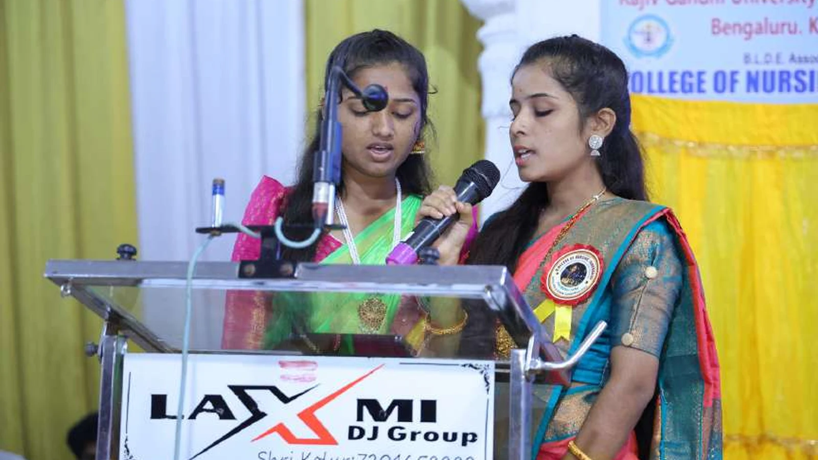 Ms Usha Pujari and Ms Kaveri Pujari performing prayer song at podium during RGUHS Kalaburagi Zone Chess Men and Women Tournament 2024 inauguration ceremony at BLDEAS College of Nursing Jamkhandi with Rajiv Gandhi University of Health Sciences College of Nursing banner visible August 23 2024
