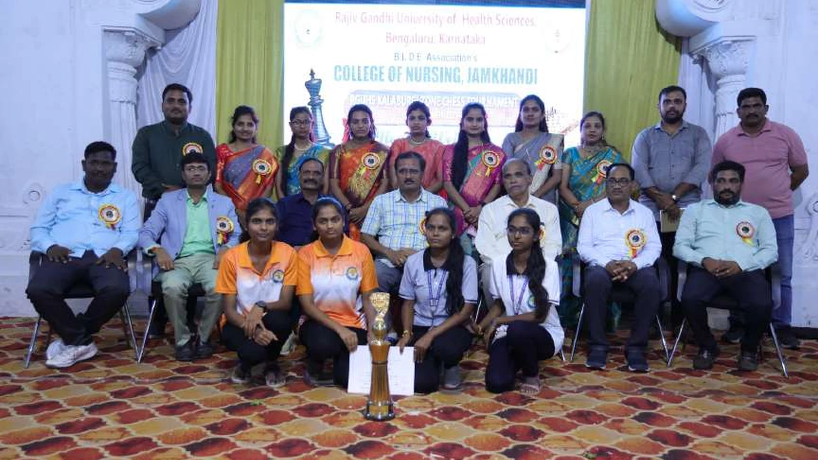 BLDEAS College of Nursing Jamkhandi women chess team fourth place winners with trophy and organizing dignitaries and staff posing together at RGUHS Kalaburagi Zone Chess Men and Women Tournament 2024 at Darbar Hall BLDEA Campus Jamkhandi August 2024 with College of Nursing Jamkhandi banner visible