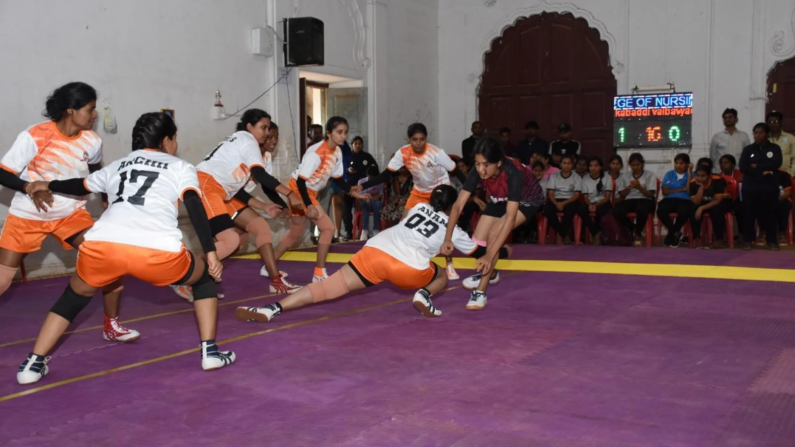 Women kabaddi players in action on purple mat during RGUHS Kabaddi Women Single Zone Tournament 2025-26 hosted by BLDEA College of Nursing Jamkhandi with digital scoreboard visible in background