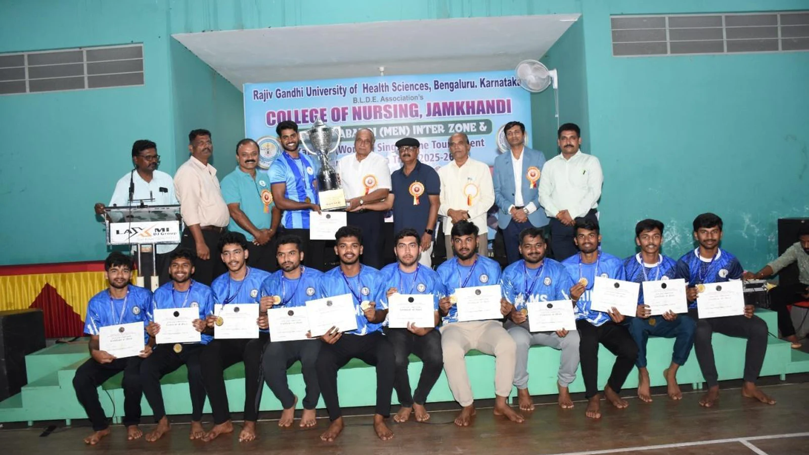 Hassan Institute of Medical Sciences Hassan Men Kabaddi team winners receiving trophy and certificates at RGUHS Kabaddi Men Inter Zone Tournament 2025-26 prize distribution function at BLDEA College of Nursing Jamkhandi