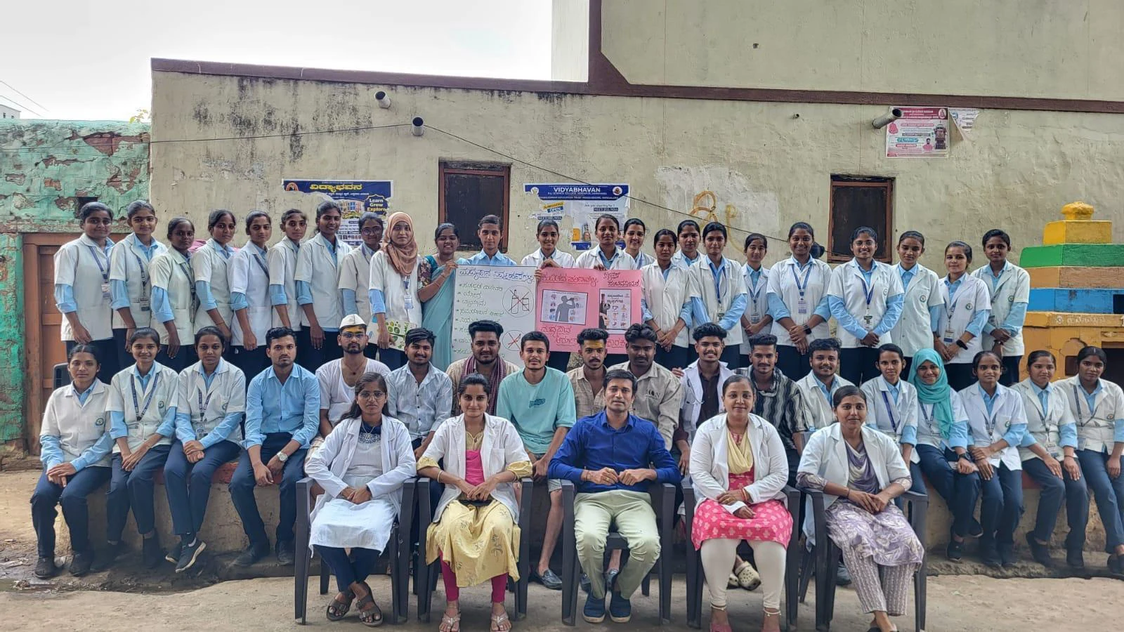 Full group photo of Seventh Semester BSc Nursing students BLDEA College of Nursing Jamkhandi with Mrs Shivaleela Sarawad and Ms Shashikala Mudhol holding health education chart after Drug Addiction Awareness Role Play at Kakanwadi Village June 2026