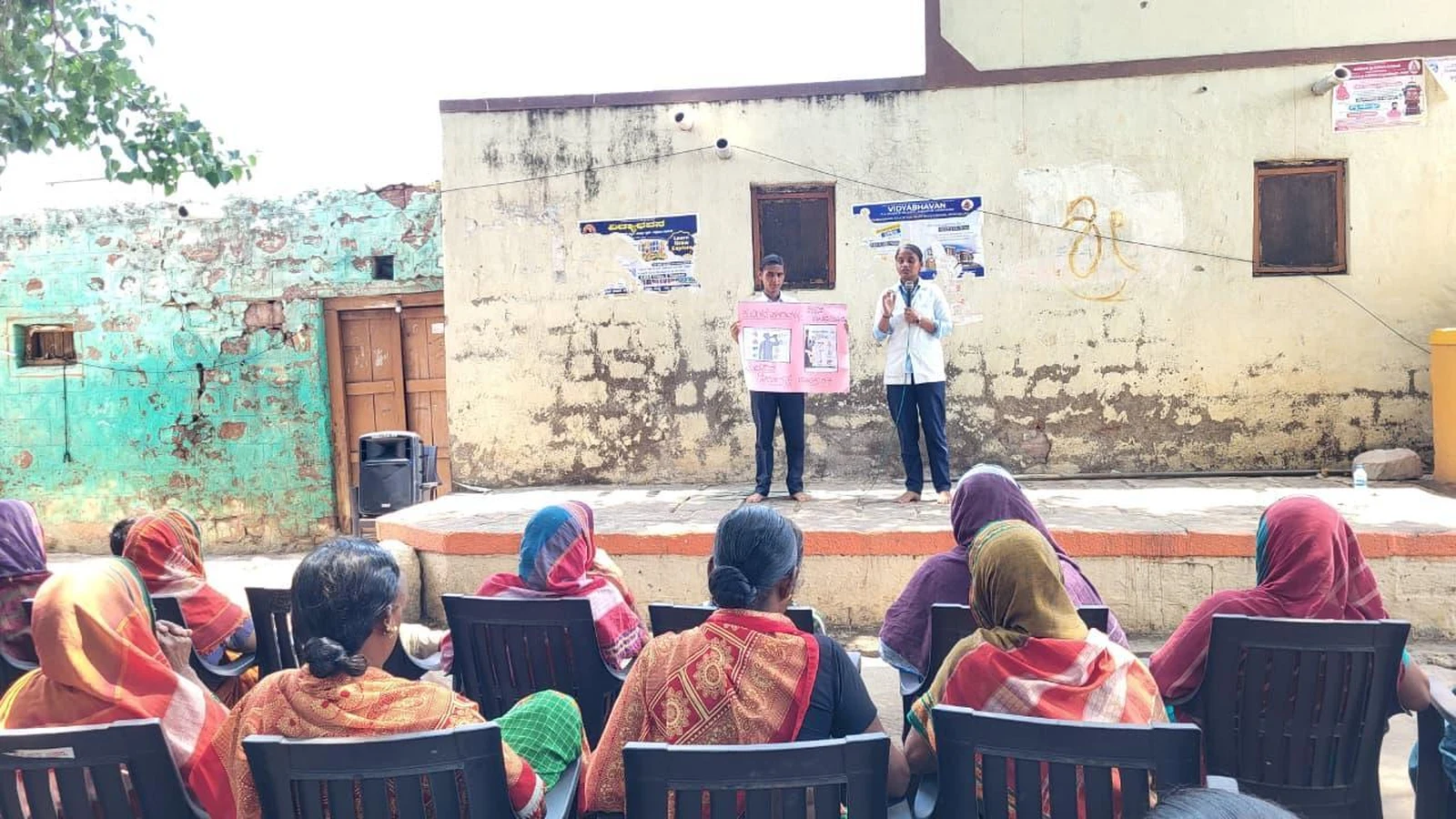 Fifth Semester BSc Nursing students BLDEA College of Nursing Jamkhandi holding pink health education chart on stage presenting Tobacco and Alcohol Prevention awareness to seated community audience at Kakanwadi Village June 2026