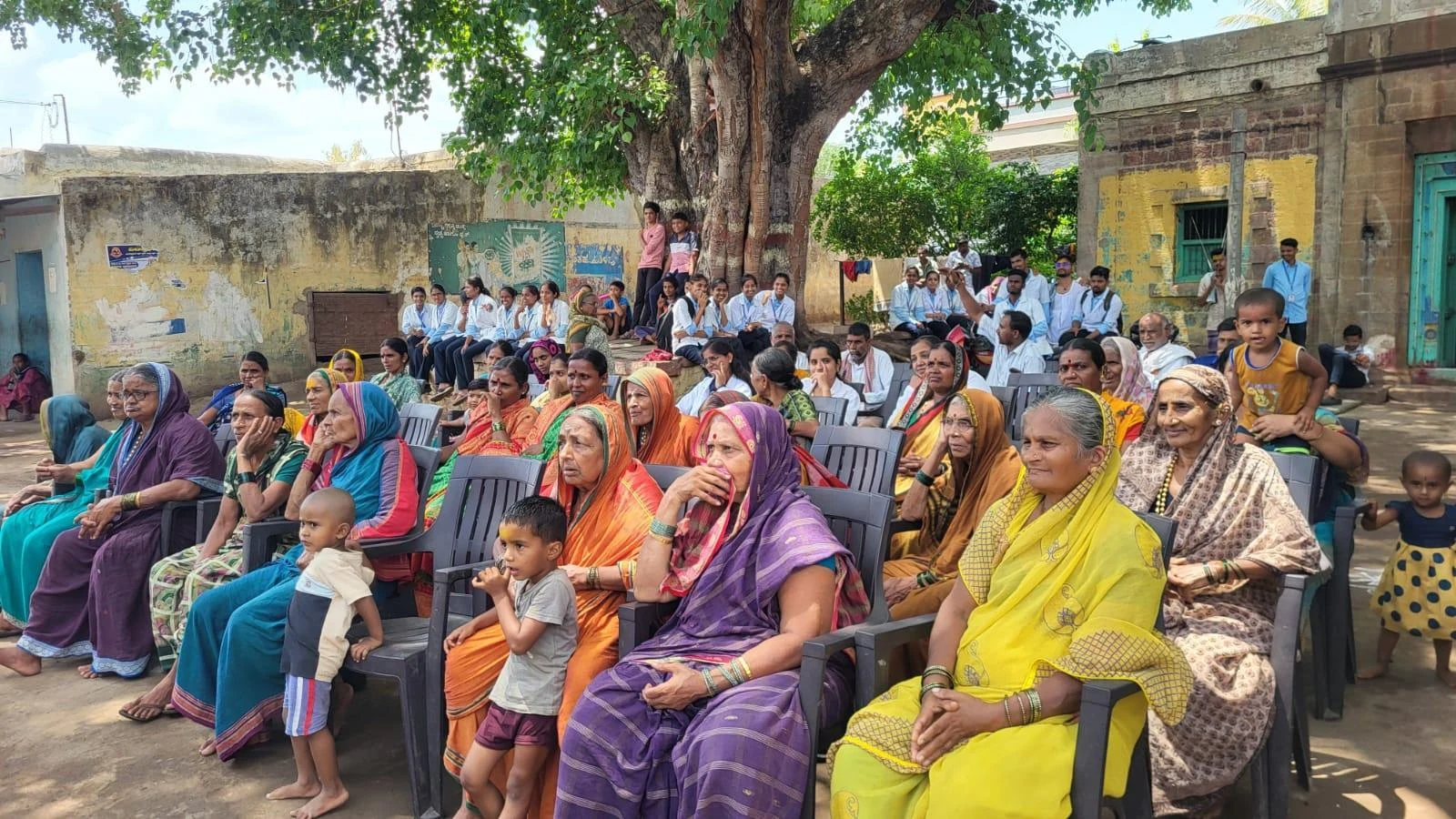 Large gathering of Kakanwadi Village community members predominantly women in colourful sarees seated outdoors under large tree watching Fifth Semester BSc Nursing students BLDEA College of Nursing Jamkhandi Tobacco and Alcohol Prevention programme June 2026