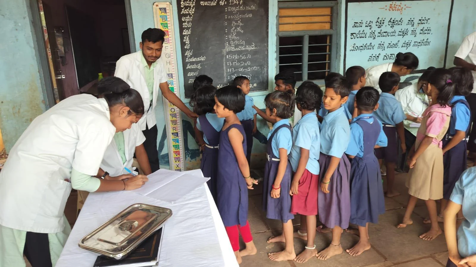 Final year BSc Nursing students from BLDEAS College of Nursing Jamkhandi measuring height of children using height chart at Government Primary School Mareguddi during School Health Program June 13 2025 with queue of school children in blue uniforms