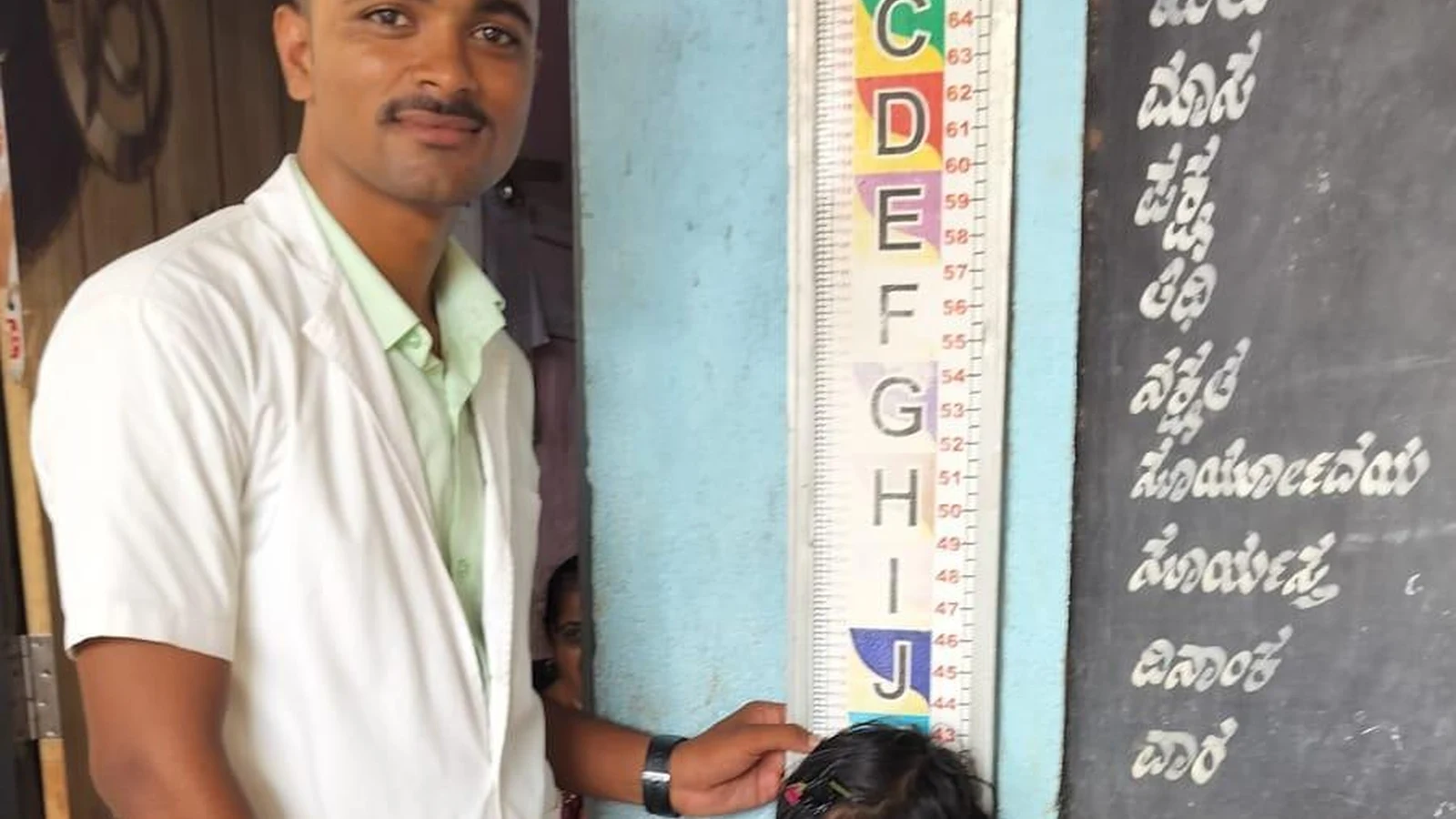 Male nursing student from BLDEAS College of Nursing Jamkhandi measuring height of a girl child using colorful height chart mounted on wall during anthropometric assessment School Health Program at Government Primary School Mareguddi June 13 2025