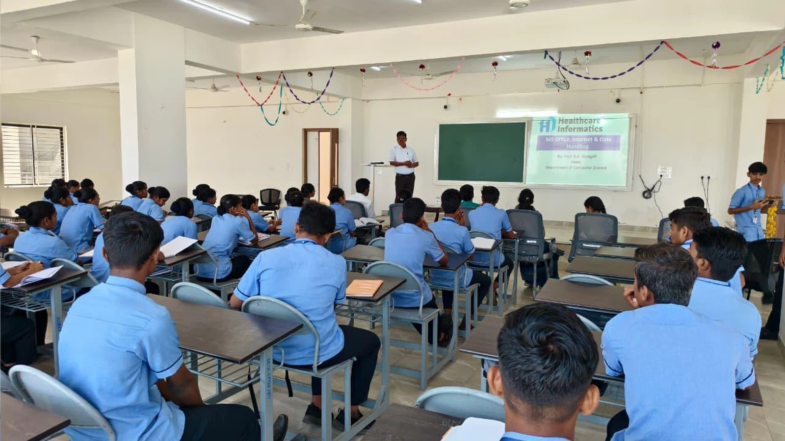 Wide view of Workshop on Digital Literacy for Health Care at BLDEA College of Nursing Jamkhandi with Second Semester BSc Nursing students in blue uniforms seated and resource person presenting at front with Health Informatics slide on projector screen November 10 2025