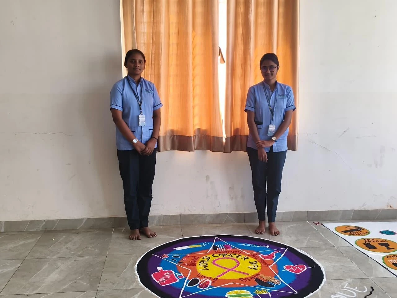 Two students standing beside their rangoli – World Cancer Day 2026, BLDEA College of Nursing Jamkhandi